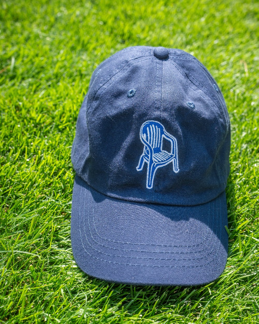 Navyblaue Cap mit gesticktem weißen Stuhlmotiv, frontal auf grünem Rasen liegend und vollständig sichtbar.
