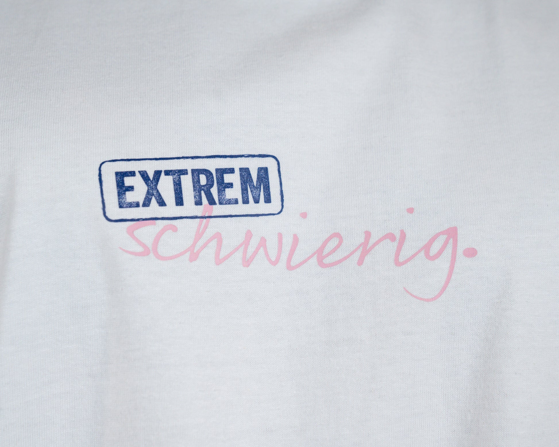 Detailaufnahme des Brustprints auf einem weißen T-Shirt. Das Wort „EXTREM“ ist in einem blauen, rechteckigen Rahmen in Großbuchstaben gedruckt, darunter steht in rosa Schreibschrift das Wort „schwierig.