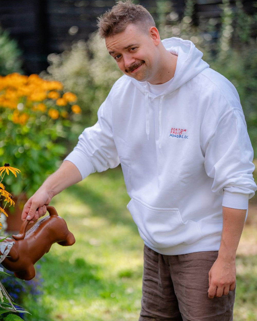 Weißer Kapuzenpullover mit kleinem Frontprint „DOKTOR FROID MONOBLOC“ in Rot und Blau. Die tragende Person gießt mit einer Gießkanne Blumen in einem Garten.