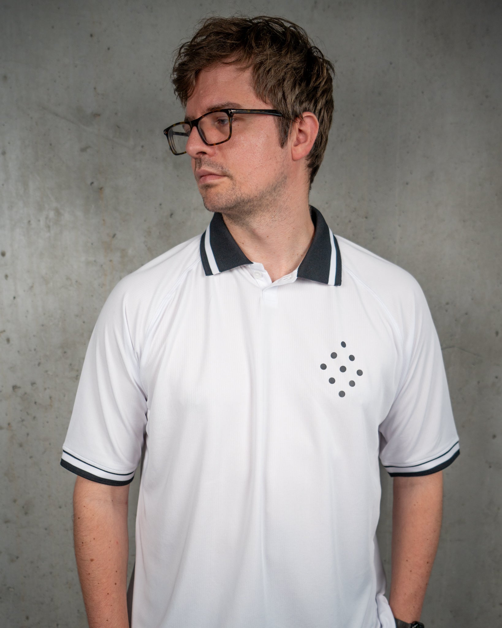 Ein Mann mit Brille trägt ein weißes Trikot mit schwarzem Polokragen und schwarzen Bündchen an den Ärmeln. Auf der linken Brustseite befindet sich ein schwarzes, rautenförmig angeordnetes Punktmuster aus neun Punkten. Der Mann steht vor einer grauen Betonwand und blickt nach links.