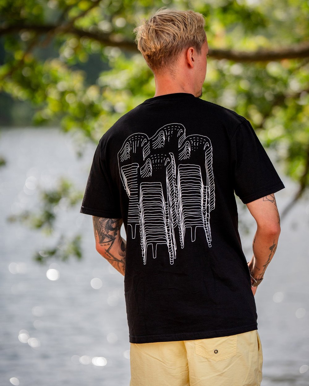 Schwarzes T-Shirt mit großem weißen Rückendruck, der mehrere gestapelte Gartenstühle in stilisierter Liniengrafik zeigt. Das Shirt wird von hinten gezeigt, getragen von einer tätowierten Person vor einem See.