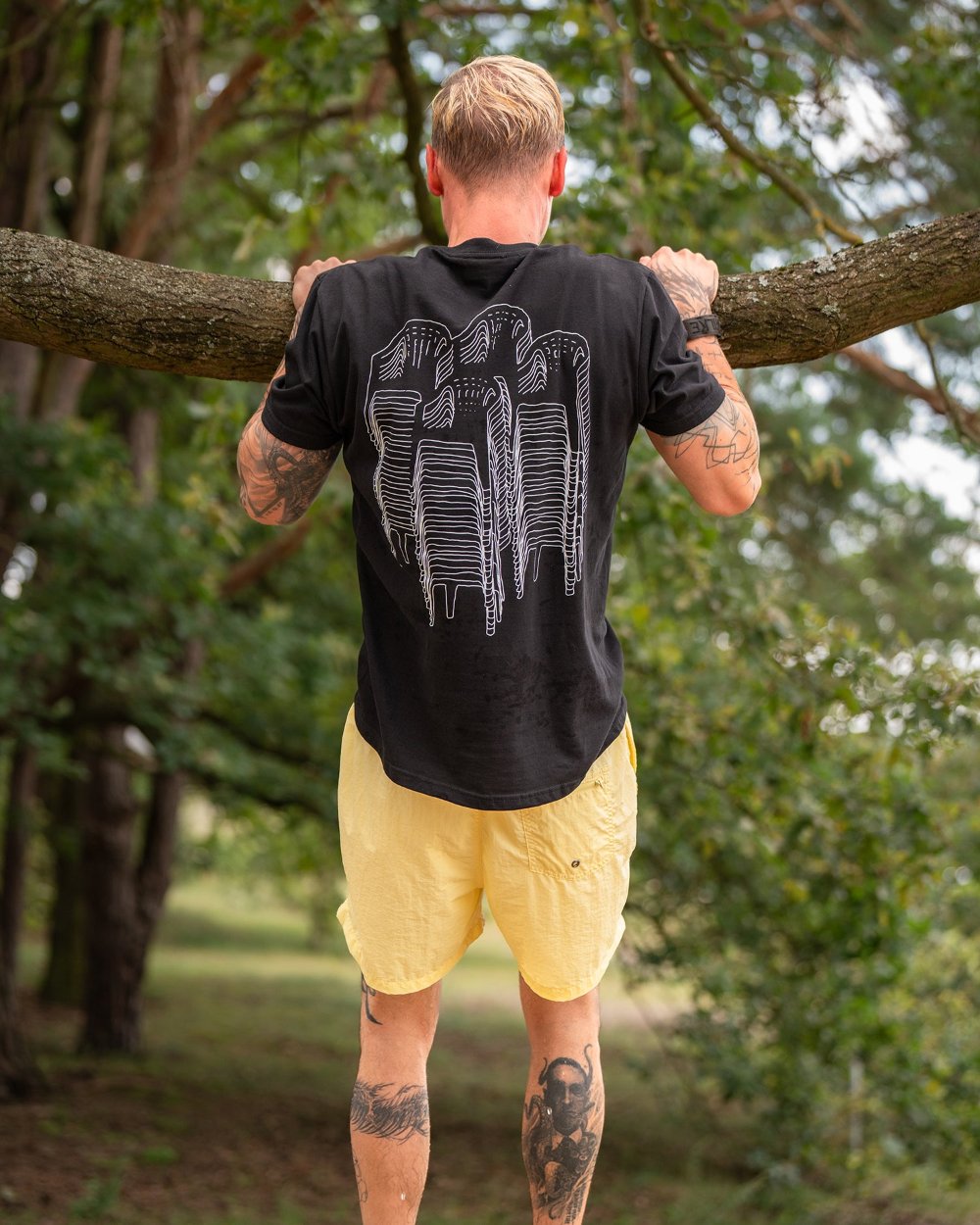 Rückansicht eines schwarzen T-Shirts mit großflächigem weißem Rückendruck, der aus übereinander gestapelten Gartenstühlen in Liniengrafik besteht. Die Person macht Klimmzüge an einem Baumast.
