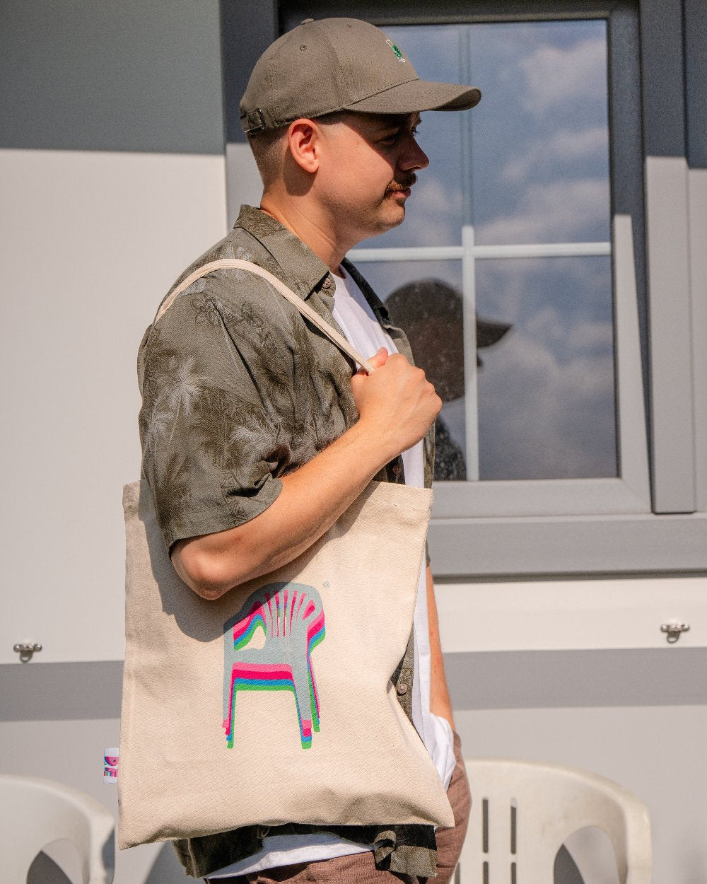 Person trägt eine beigefarbene Stofftasche mit breiten Trägern über der Schulter. Auf der Vorderseite ist ein stilisiertes Monobloc-Stuhl-Motiv in pink, türkis und grünem Versatzdruck abgebildet.