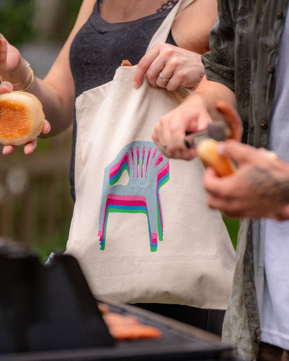 Nahaufnahme der Stofftasche während eines Grillfestes. Die Tasche zeigt ein grafisches Stuhlmotiv in mehrfarbigem Versatzdruck (pink, türkis, grün). Die Tasche wird lässig über der Schulter getragen.