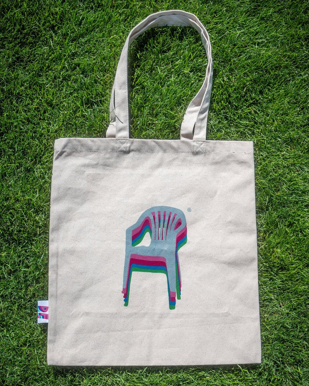 Beigefarbene Stofftasche mit breiten Trägern liegt flach auf grünem Rasen. Zentral auf der Vorderseite ist ein bunter Versatzdruck eines Monobloc-Stuhls in grau, pink, türkis und grün zu sehen.