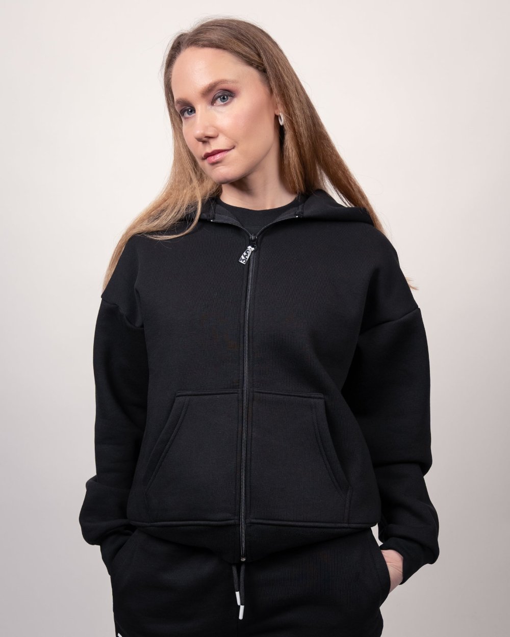 Eine Frau trägt einen schwarzen Zip-Hoodie mit Kapuze. Die Jacke ist geschlossen, hat eine Kängurutasche vorne und zeigt keine sichtbaren Logos auf der Vorderseite.
