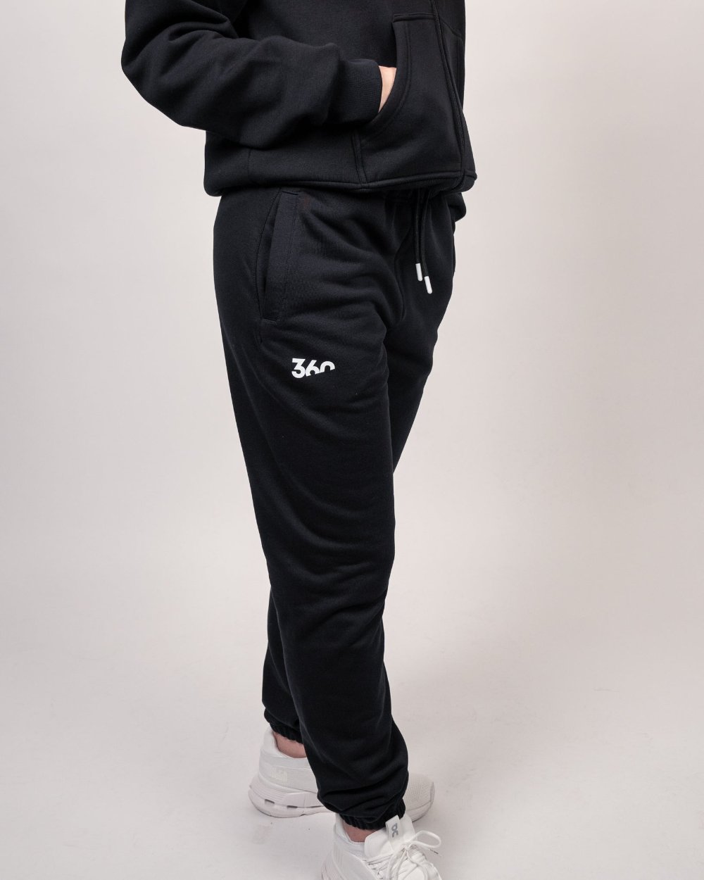 Schwarze Jogginghose mit elastischem Beinabschluss und weißem „360“-Logo auf dem linken Oberschenkel, getragen zu weißen Sneakern und schwarzem Zip-Hoodie.