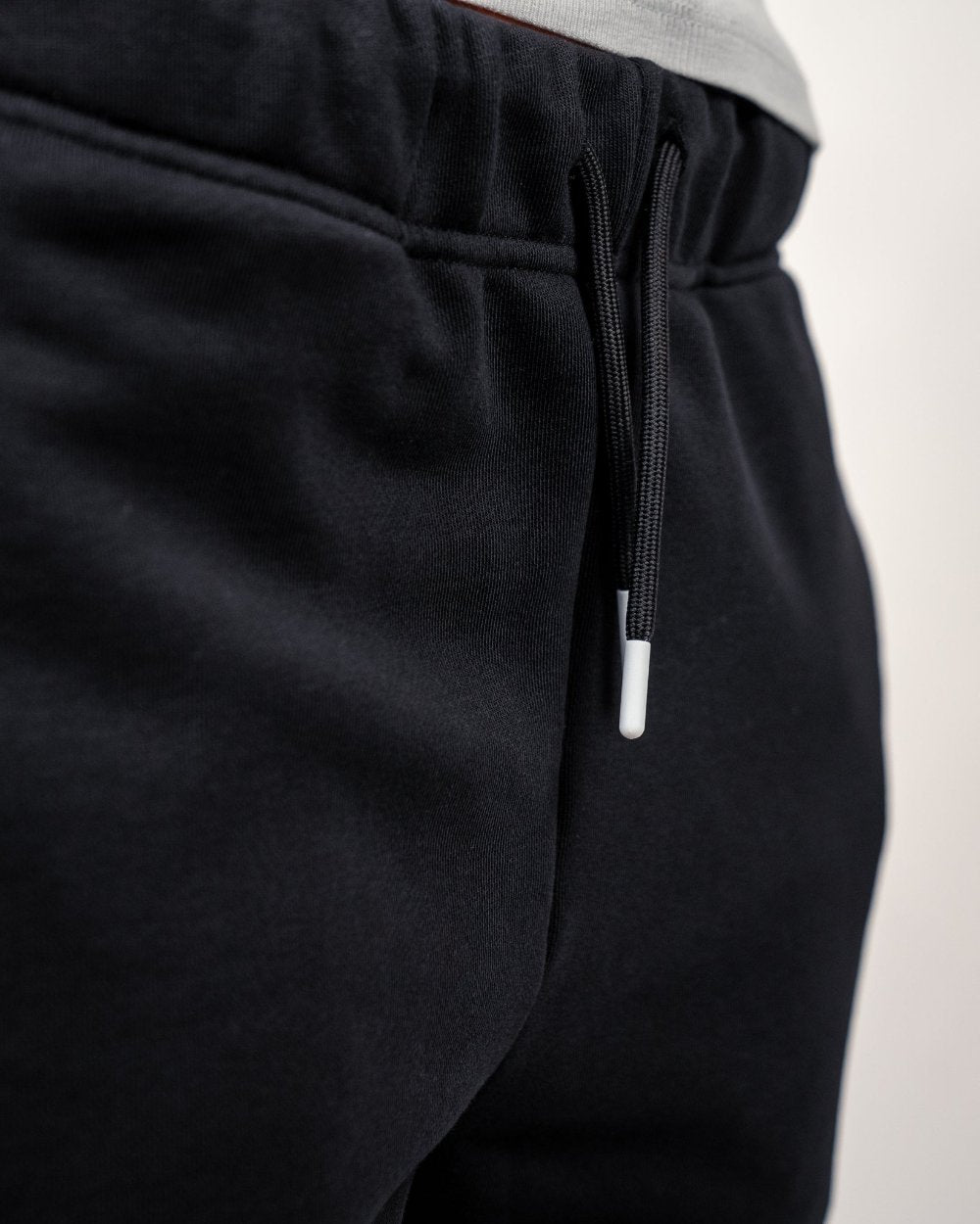 Detailansicht des Hosenbunds mit schwarzem Kordelzug und weißen Endkappen an der schwarzen Jogginghose.