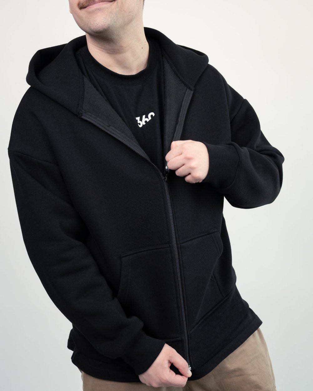 Ein Mann öffnet seinen schwarzen Zip-Hoodie. Darunter ist ein schwarzes T-Shirt mit kleinem weißen „360“-Logo zu sehen.