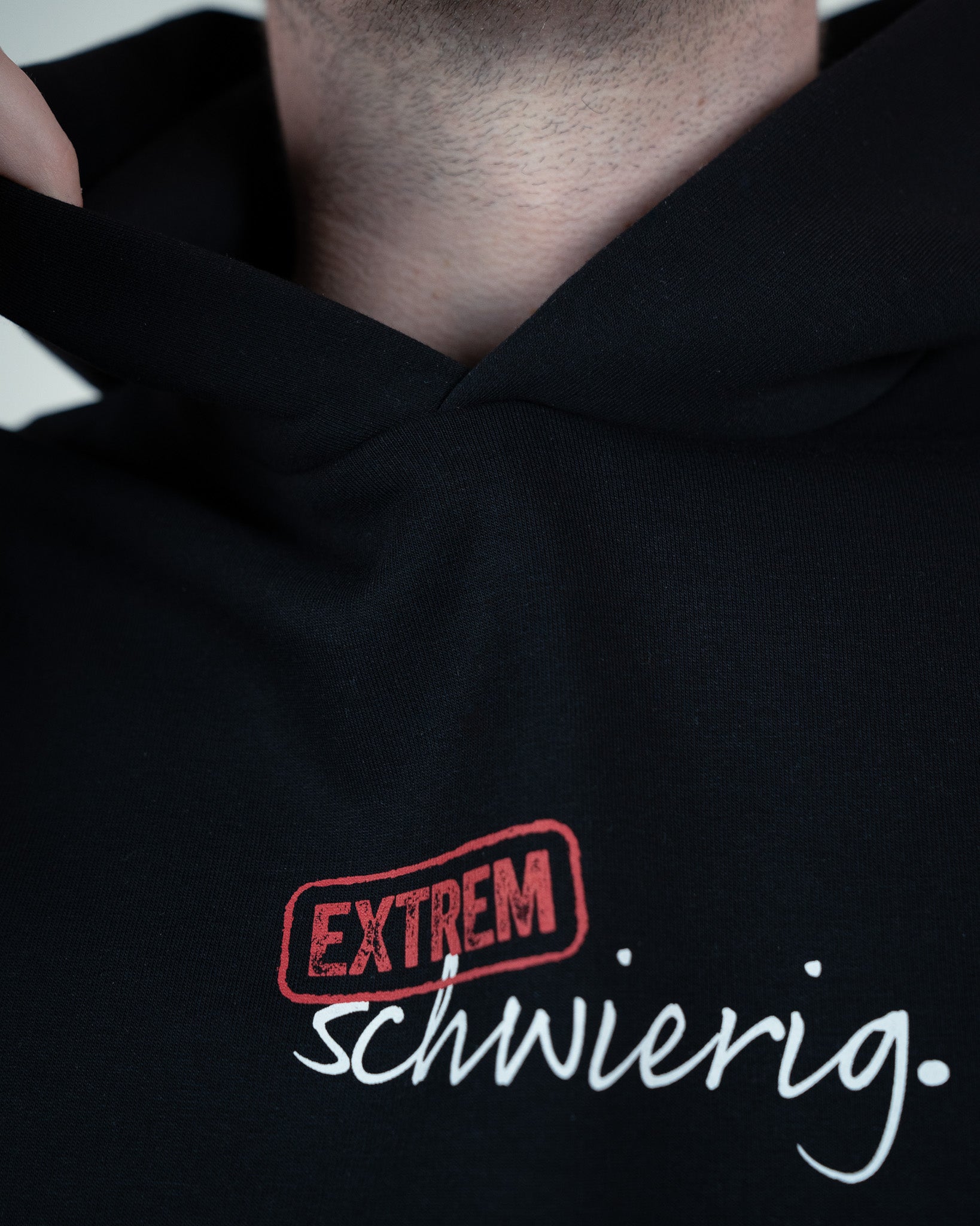 Detaillierte Aufnahme des roten „EXTREM“-Stempels und des weißen Schriftzugs „schwierig.“ auf schwarzem Stoff. Fokus auf Textur und Naht der Kapuze.