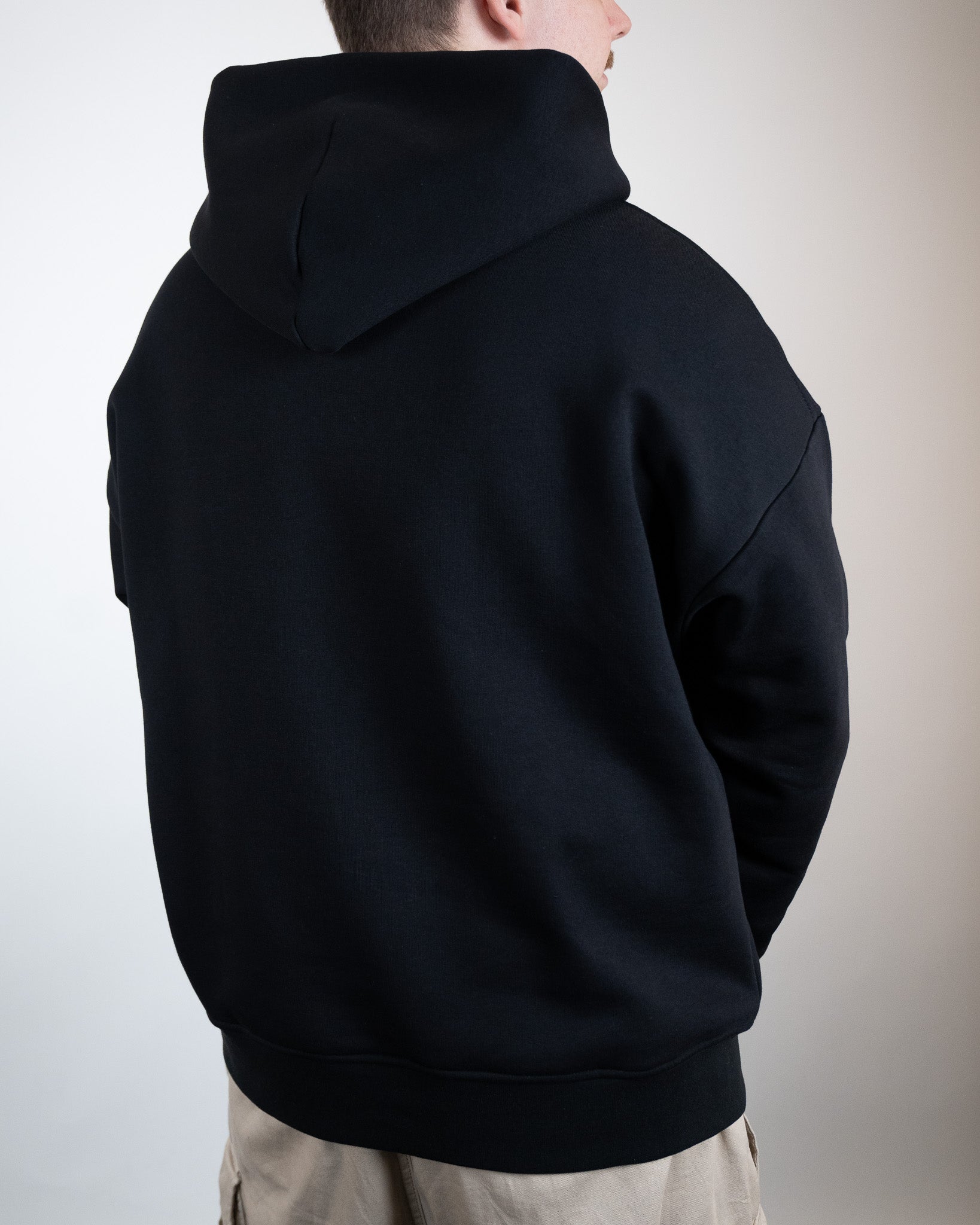 Schwarzer Kapuzenpullover von hinten leicht seitlich gezeigt. Die Kapuze ist hochgestellt, der Rücken bleibt unbedruckt.