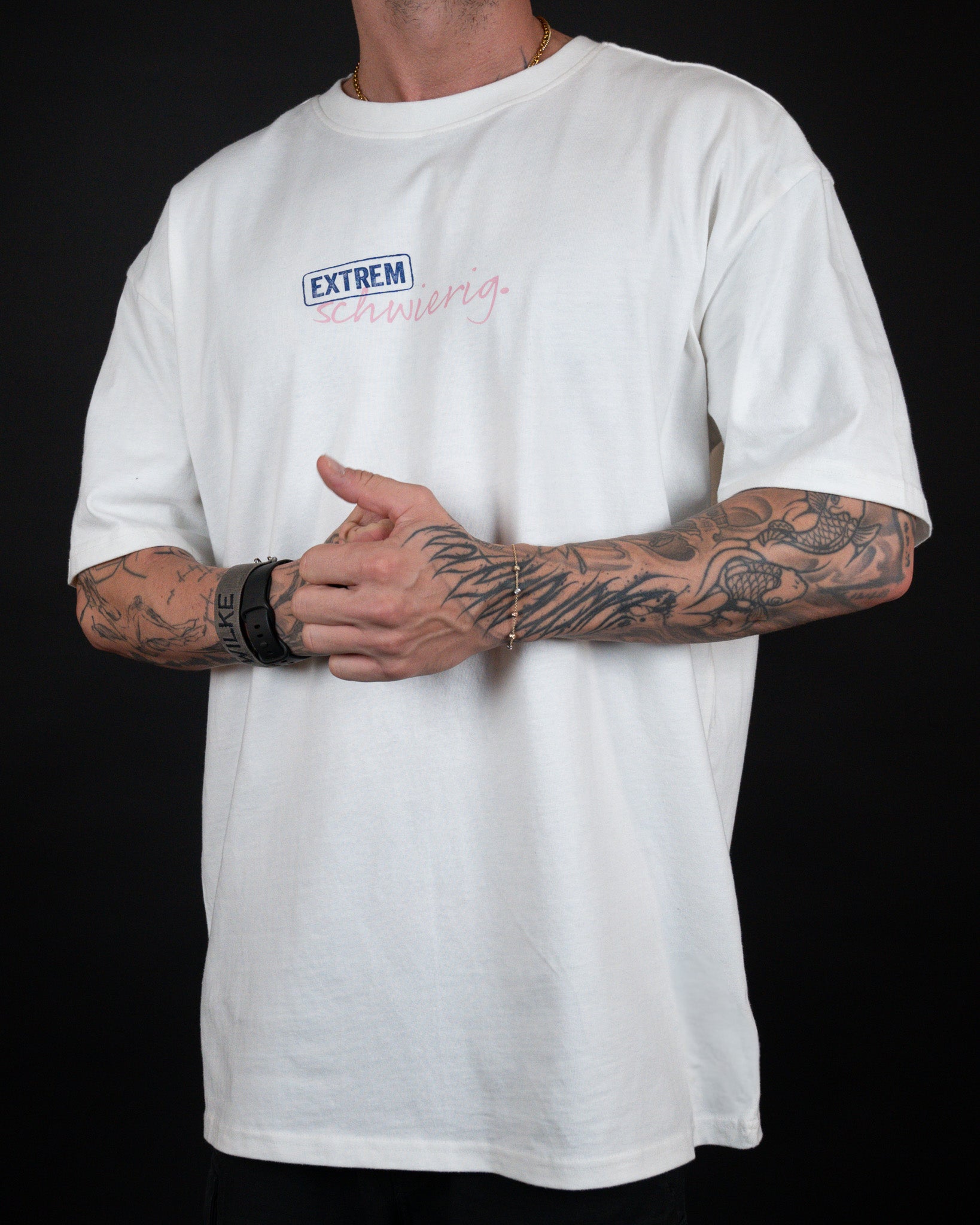 Vorderansicht eines weißen T-Shirts mit dem Schriftzug „EXTREM schwierig.“ auf der linken Brust. Das Shirt hat eine lockere Passform und wird von einer tätowierten Person getragen, die die Hände vor dem Bauch verschränkt.