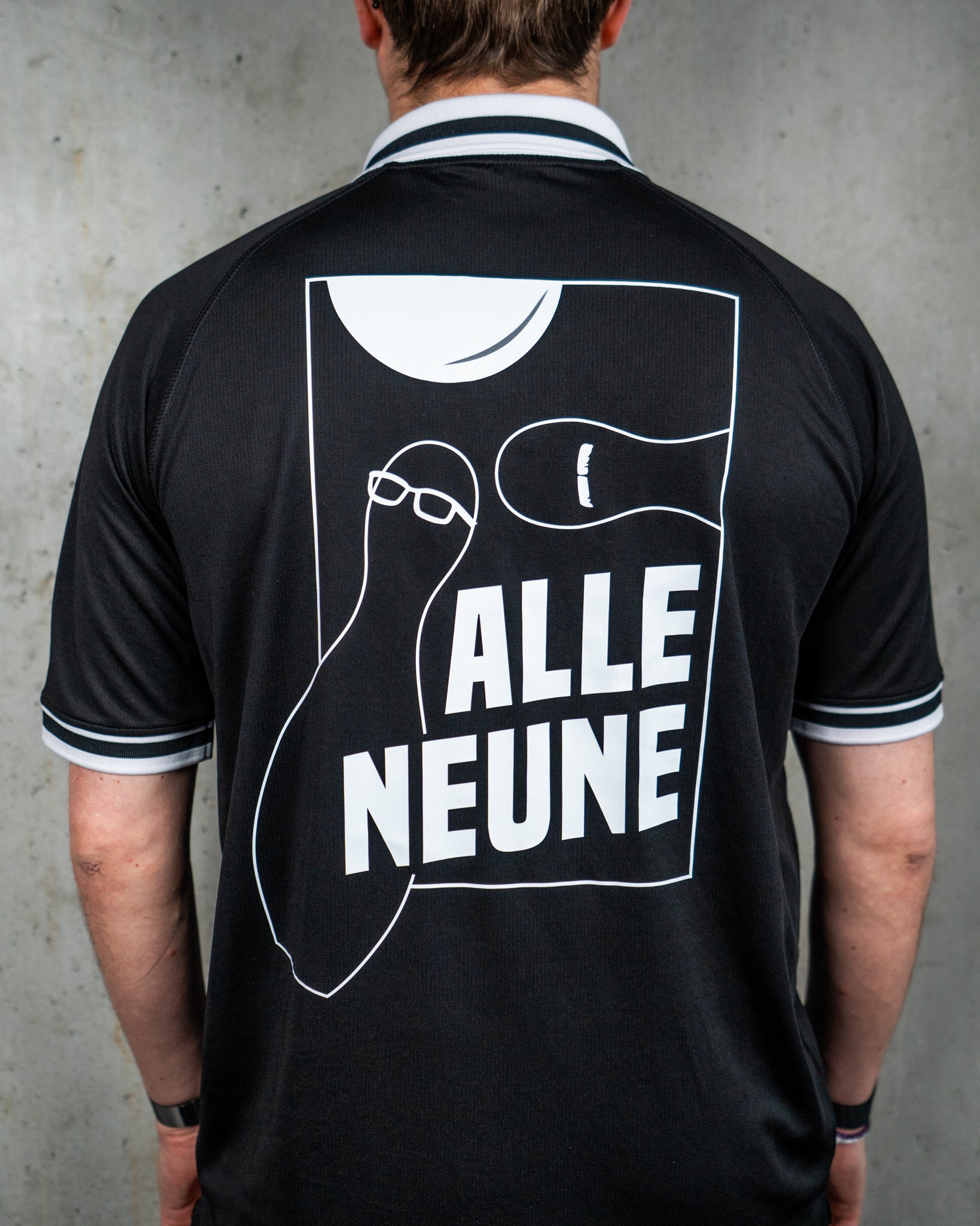 Rückenansicht eines schwarzen Trikots mit weißem Grafikprint. Das Motiv zeigt stilisierte Kegelfiguren mit minimalistischen Gesichtern – eine trägt eine Brille, die andere einen Schnurrbart. Oberhalb ist ein angedeuteter Bowlingball zu sehen. Die Szene ist in ein rechteckiges Feld eingebettet, darunter steht der Schriftzug „ALLE NEUNE“ in weißer Versal-Schrift. Das Trikot hat einen weißen Polokragen mit schwarzen Streifen sowie passende Bündchen an den Ärmeln. Der Hintergrund ist eine glatte Betonwand.




