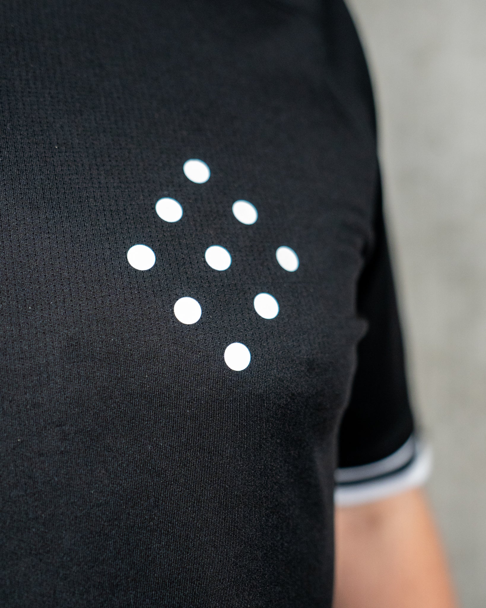 Nahaufnahme der Brustpartie eines schwarzen Trikots mit strukturierter Oberfläche. Im Fokus steht ein minimalistisches, aufgedrucktes Motiv aus neun weißen Punkten, die in Form einer Raute (4–3–2) angeordnet sind. Der Stoff zeigt eine dezente Wabenstruktur. Am rechten Bildrand ist ein Teil des weißen Ärmelbündchens mit schwarzem Streifen sichtbar.