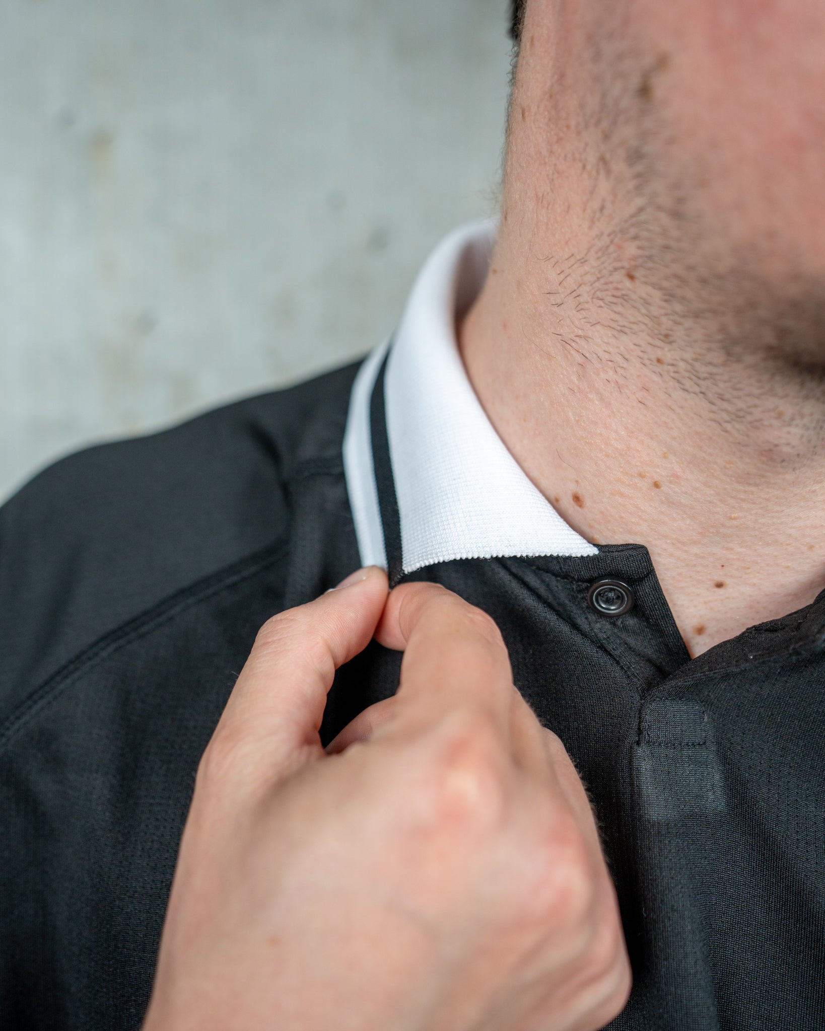 Nahaufnahme eines schwarzen Trikots mit weißen Polokragen. Der Kragen ist mit einem Schwarzen Streifen verziert. Sichtbar sind außerdem die weiße Knopfleiste und feine vertikale Strukturen im Stoff. Im Bildausschnitt sind Hals und Haaransatz einer Person zu sehen.









