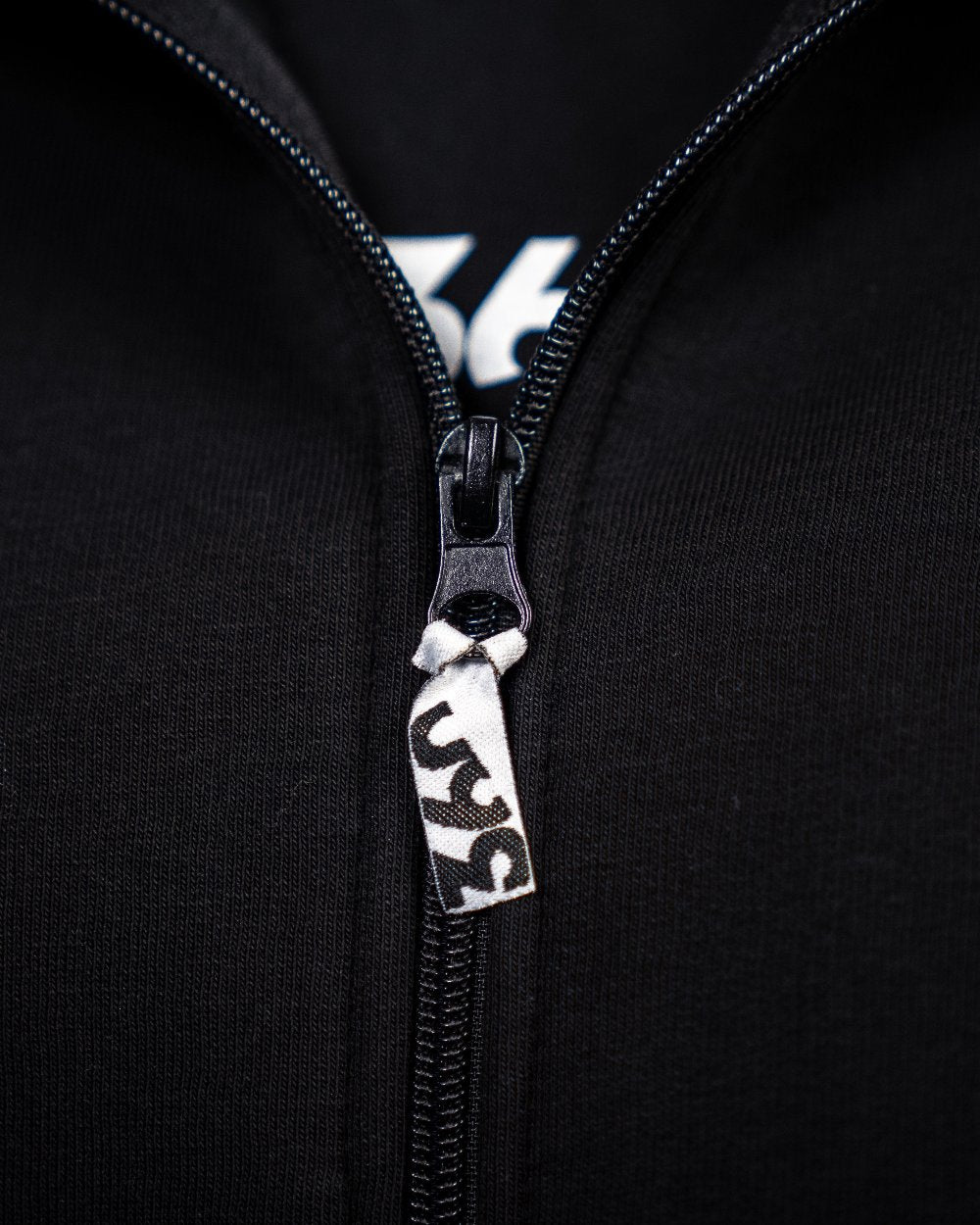 Detailaufnahme des Zippers eines schwarzen Zip-Hoodies. Am Reißverschlussanhänger ist ein kleiner Stofftag mit „360“-Logo befestigt.