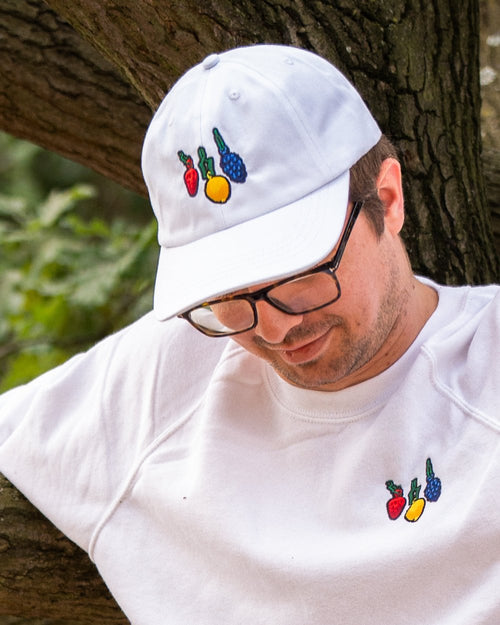 Nahaufnahme einer weißen Baseballcap mit gestickten Fruchtmotiven auf der Vorderseite. Die Stickerei zeigt von links nach rechts eine rote Erdbeere, eine gelbe Zitrone und eine blaue Traube, alle mit grünem Stiel. Die Cap wird von einer Person mit weißem Sweatshirt getragen, das dieselbe Fruchtstickerei auf der Brust trägt.
