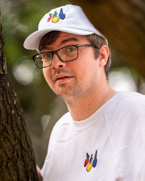 Porträtaufnahme einer Person mit weißer Cap und passendem weißen Sweatshirt, beide mit identischer Fruchtstickerei. Die Stickerei auf der Cap ist gut sichtbar und zeigt eine Erdbeere, eine Zitrone und eine Traube mit grünem Stiel.