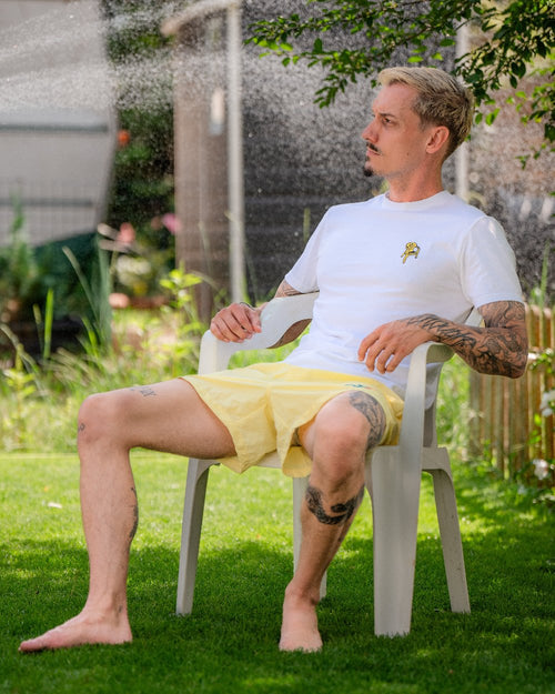 Der Mann sitzt barfuß und entspannt auf einem weißen Plastikstuhl im Garten. Er trägt ein weißes T-Shirt und eine gelbe Badeshorts mit sichtbarem gesticktem Logo auf dem linken Bein. Im Hintergrund sprüht Wasser aus einem Rasensprenger.